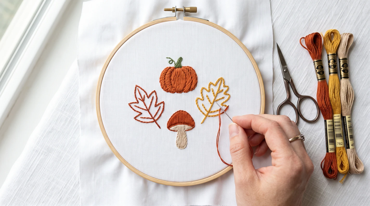 Autumn hoop with a small orange pumpkin, mushroom and two leaf outlines beside warm-colored floss