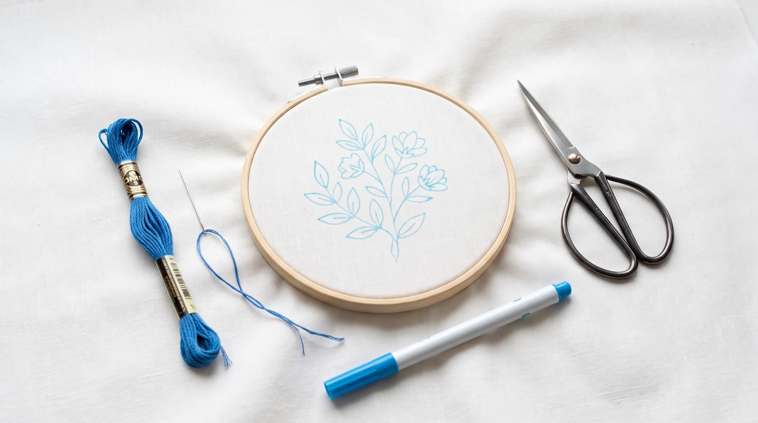 Smooth white fabric in a hoop with a blue botanical outline beside floss, needle, scissors and marking pen