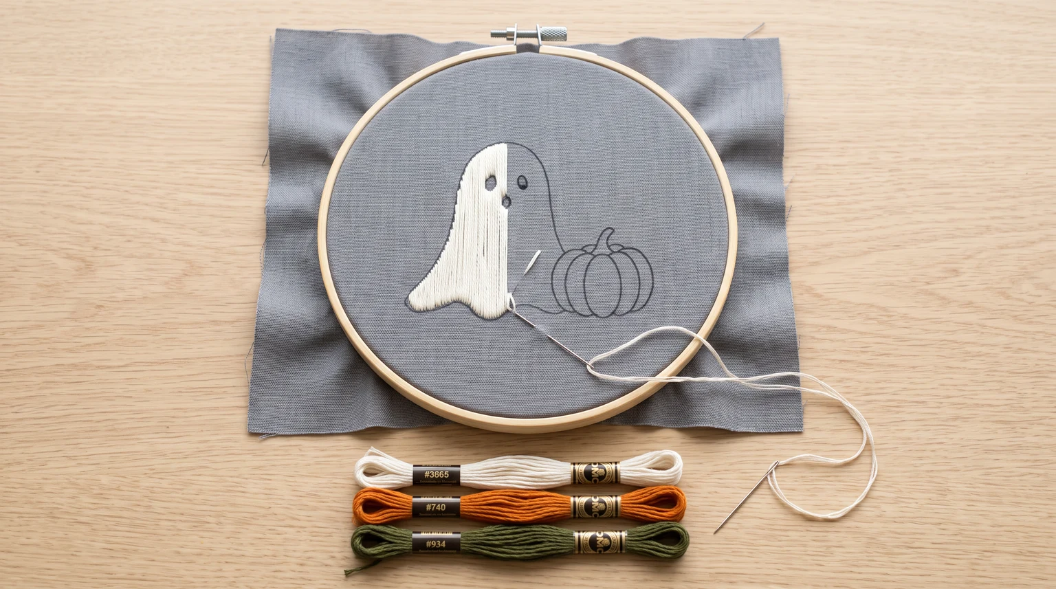 Small white embroidered ghost beside a pumpkin outline on gray fabric in a hoop