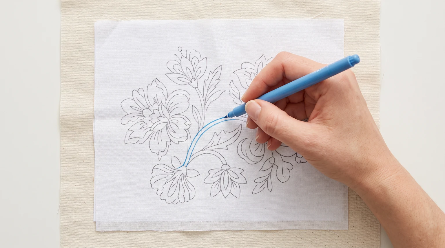 Hand using a blue marking tool to trace a botanical flower outline through light fabric